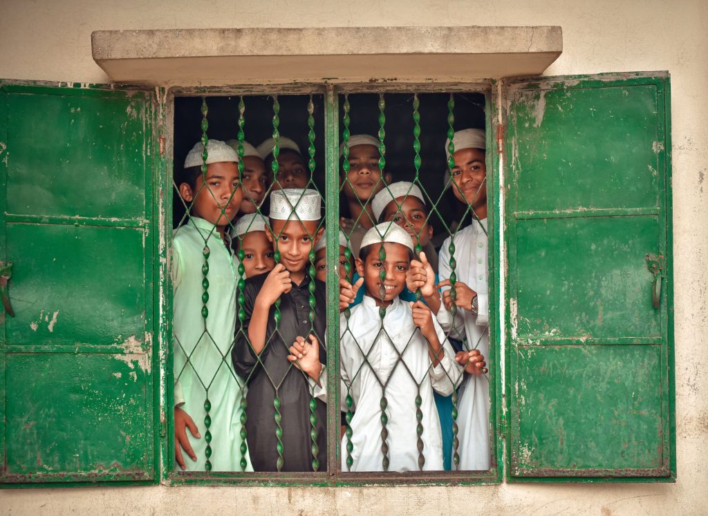 A group of boys in Islamic attire looking through a window with green shutters, embodying cultural attire and togetherness.