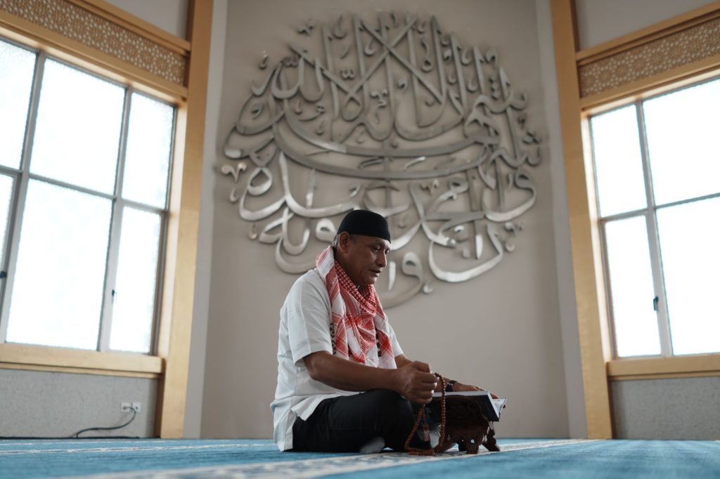A man in traditional attire reading Quran inside a mosque in Indonesia, adorned with intricate calligraphy.