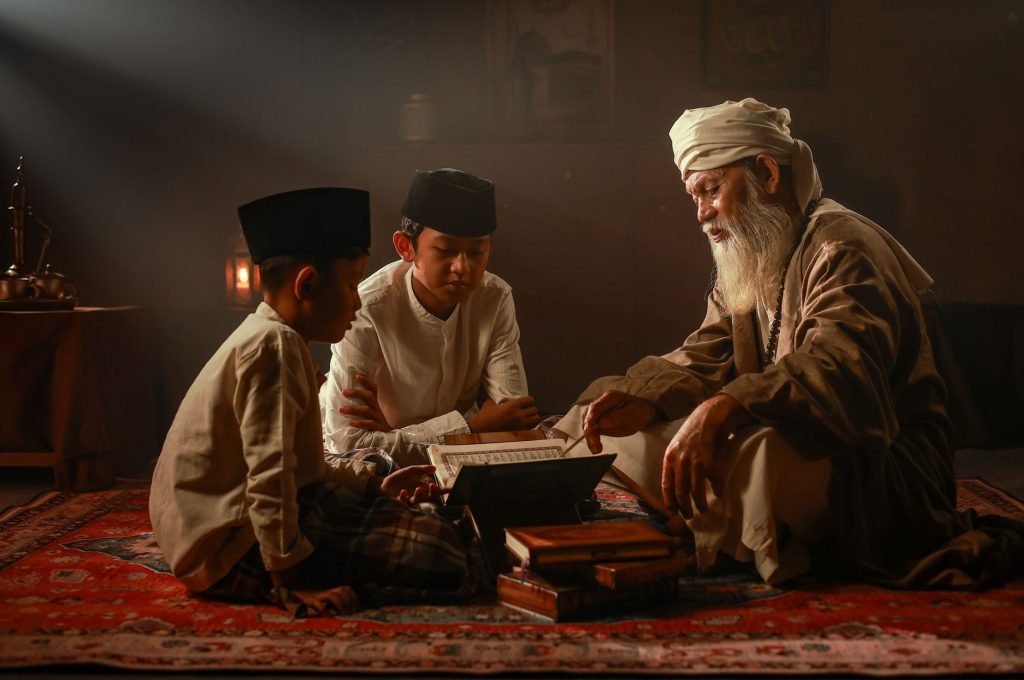 Elderly man teaching two boys in traditional Islamic attire indoors in Jakarta.