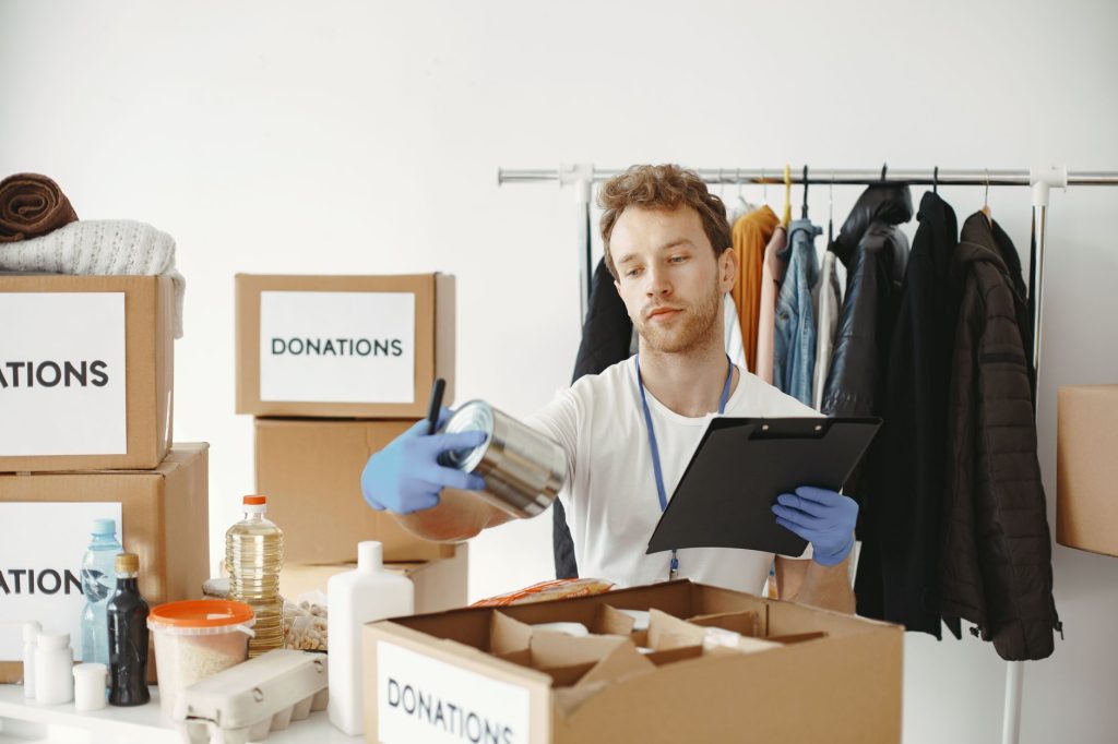 A volunteer organizes food and clothing donations in a community center, maintaining records on a clipboard.