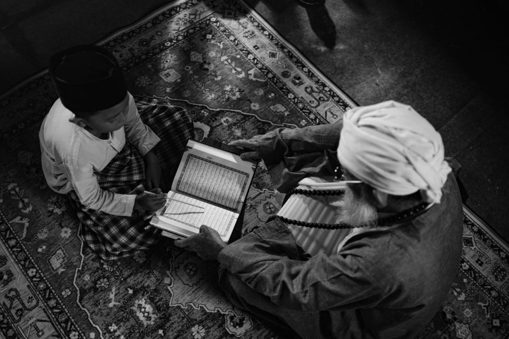 Black and white image of a boy learning from an elder, highlighting traditional Islamic education in Jakarta.